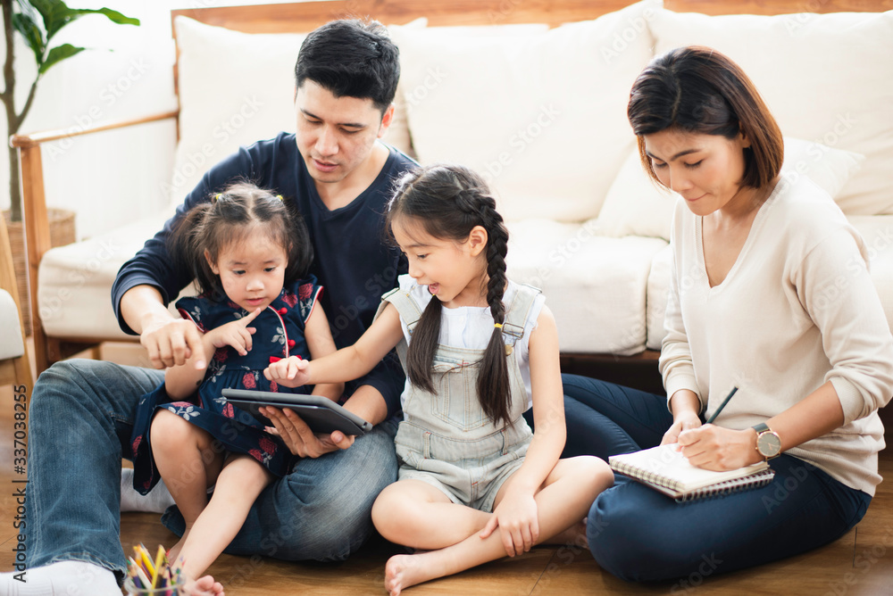 Young Asian love family stay in living room at home, father teaching two daughter to playing and use the tablet by looking which sit on wooden floor near sofa, looking smiling and felling happy. Young Asian love family stay in living room at home, father teaching two daughter to playing and use the tablet by looking which sit on wooden floor near sofa, looking smiling and felling happy.
