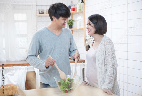 Asian man and pregnancy woman stand in kitchen at home.Father stand and look mother tummy mix salad feeling love expecting about future newborn baby. Love and care of pregnant mother   s day concept.