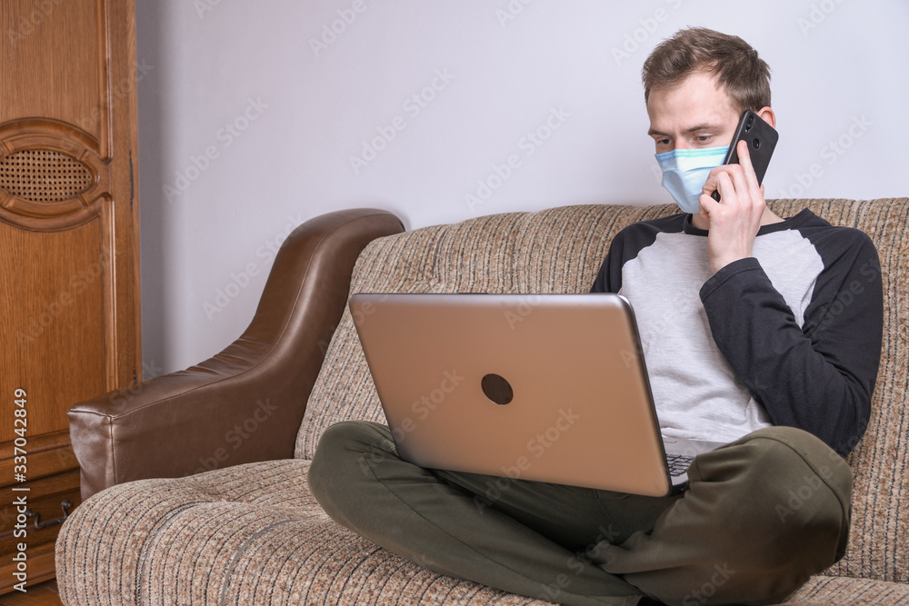 Young man in medical mask working at home in the room on the sofa using a laptop. Quarantine, self-isolation, coronavirus protection. Vacation from work. Young man in medical mask working at home in the room on the sofa using a laptop. Quarantine, self-isolation, coronavirus protection. Vacation from work.