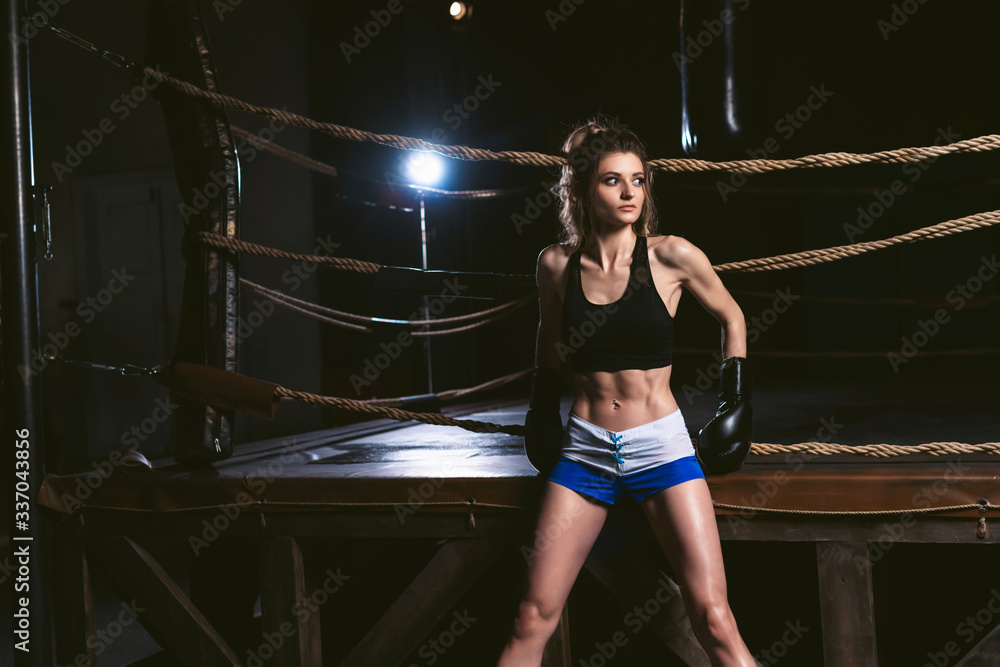 Young girl in sportswear: a t-shirt and shorts wearing boxing gloves. Athletic woman posing on a background of a boxing ring in the dark gym. Power and motivation concept Young girl in sportswear: a t-shirt and shorts wearing boxing gloves. Athletic woman posing on a background of a boxing ring in the dark gym. Power and motivation concept