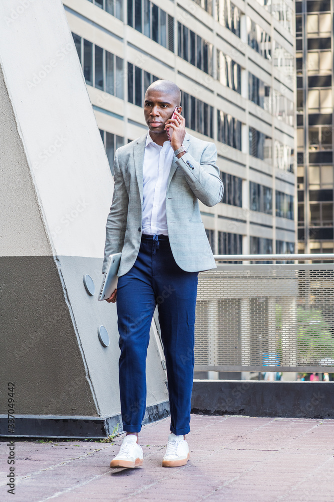 African American Man traveling, working in New York wearing blazer, blue pants, white sneakers, carrying laptop computer, walking outside office building, talking on cell phone,. Stock Photo | Adobe