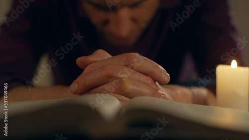 A man is sitting praying fervently at night with hands together in front of an open Bible and a candle light