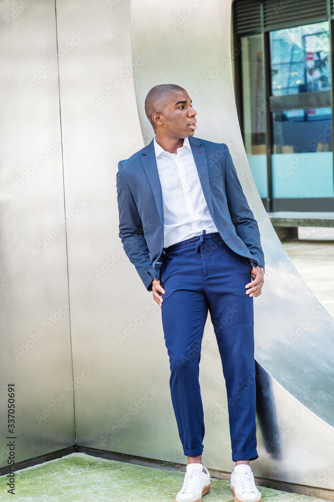 Young African American college student relaxing, waiting outside in New York City, wearing blue blazer, white shirt, blue pants, white sneakers, standing against silver metal wall, looking away.. Young African American college student relaxing, waiting outside in New York City, wearing blue blazer, white shirt, blue pants, white sneakers, standing against silver metal wall, looking away..