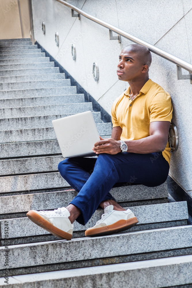 Young African American Man working in New York City, wearing yellow shirt, blue pants, white sneakers, sitting on stairs outside office building, working on laptop computer, looking up, thinking..