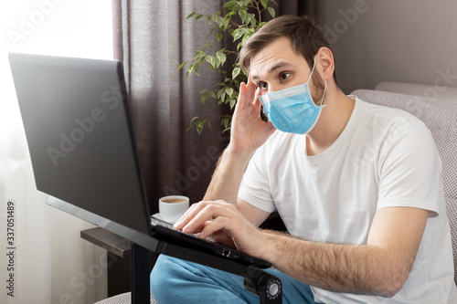 Young caucasian gloomy man in stress working from home office wearing protective mask using laptop and internet. Coronavirus covid 19 quarantine. Remote work, freelancer, home office workplace on sofa