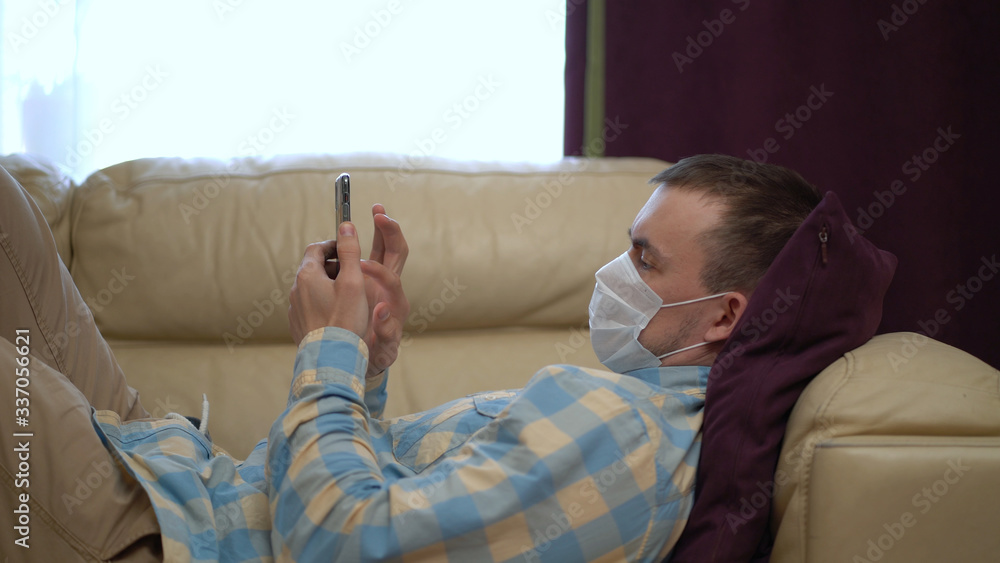Man in a Medical Mask Uses a Smartphone at Home During Quarantine. Works Alone From Home Remotely. Quarantine Covid-19 Corona Virus. Social Distancing Concept. Man in a Medical Mask Uses a Smartphone at Home During Quarantine. Works Alone From Home Remotely. Quarantine Covid-19 Corona Virus. Social Distancing Concept.