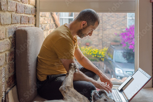 Young man in sweat pants and t-shirt typing in his laptop while his dog ask for attention. Home office concept © Julian