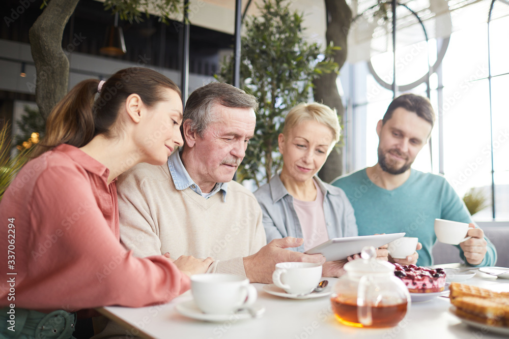 Mature man holding digital tablet in his hands and showing something online to his family while they sitting in cafe Mature man holding digital tablet in his hands and showing something online to his family while they sitting in cafe