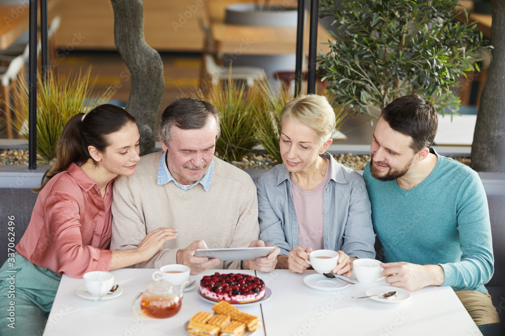 Mature man watching something on tablet pc while sitting at the table together with his family during breakfast in cafe Mature man watching something on tablet pc while sitting at the table together with his family during breakfast in cafe