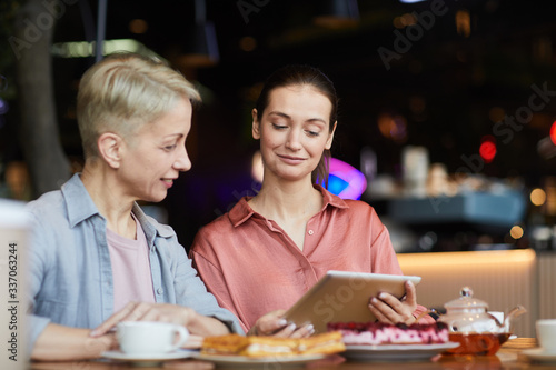 Young woman with digital tablet showing something to mature woman while they sitting at the table with tea in cafe