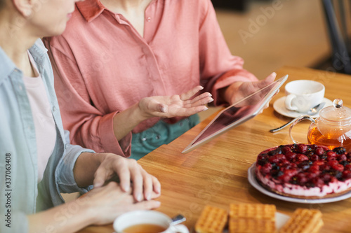Close-up of two women using digital tablet while sitting at the table and drinking tea with fruit cake