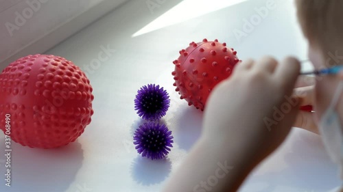 a boy in the house on the windowsill, in a medical mask and glasses, through a toy medical magnifying glass carefully examines balls with spikes of red, blue, pink color similar to a coronavirus photo