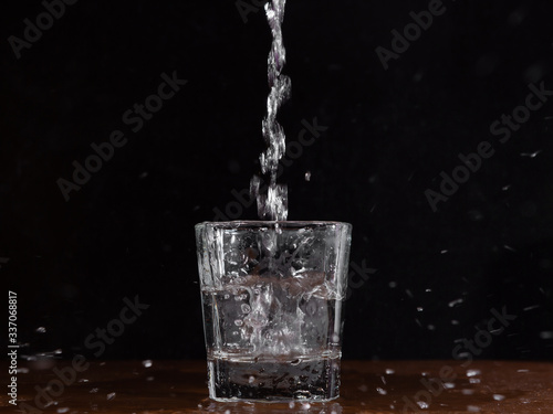A glass of water on a black background, which is filled with a jet of water and flying spray