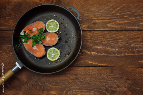 Cut pieces of raw red fish salmon in a cast iron pan with lemon slices spices and green parsley sprigs on a brown wooden background top view with an empty space for text, copy space, flat lay