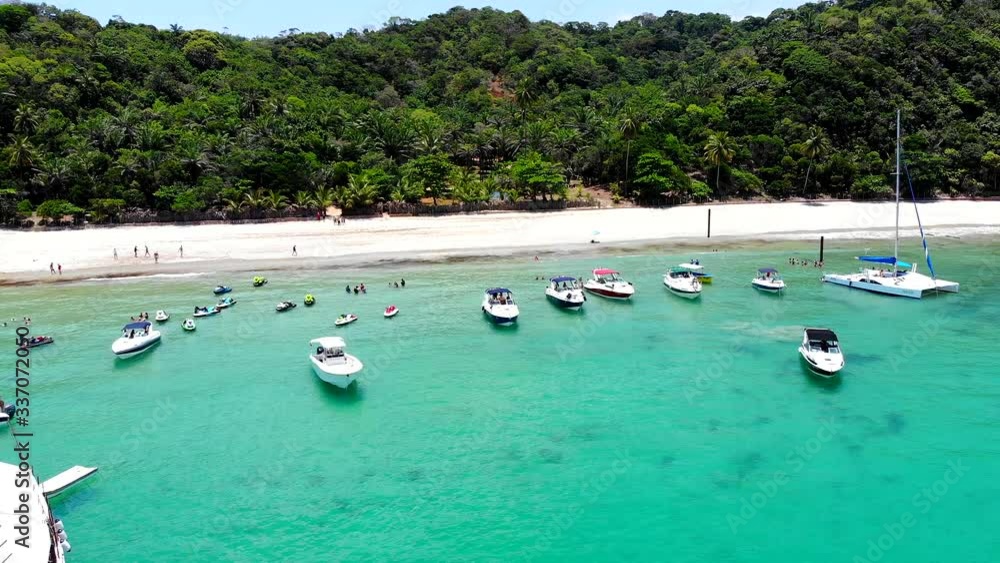 Aerial of beautiful turquoise beach with motor boats and people enjoying the sun in front of a tropical forest. Drone slowly moves forwards