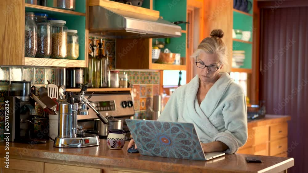 Mature woman in bathrobe, studies what is on a laptop screen and then smiles and picks up her phone and starts dialing. Mature woman in bathrobe, studies what is on a laptop screen and then smiles and picks up her phone and starts dialing.
