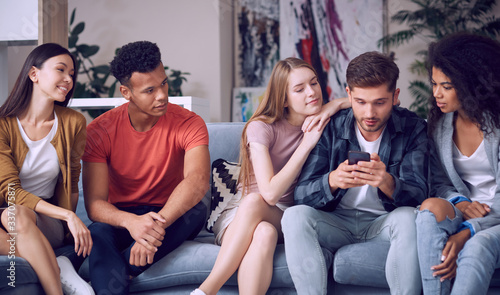 Looking at the photos. Group of young happy multicultural people in casual wear looking at smartphone, enjoying time together while sitting on the sofa in the living room