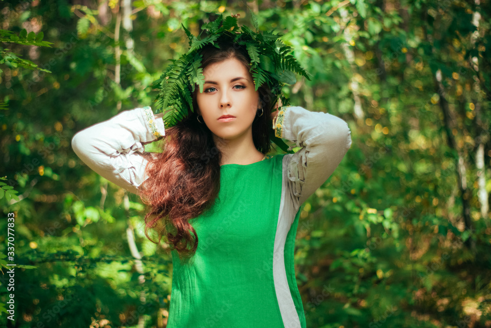 An attractive country girl in a green medieval dress with a wreath of fern on her head stands in the forest. Young witch, herbalist collects herbs for a potion. An attractive country girl in a green medieval dress with a wreath of fern on her head stands in the forest. Young witch, herbalist collects herbs for a potion.