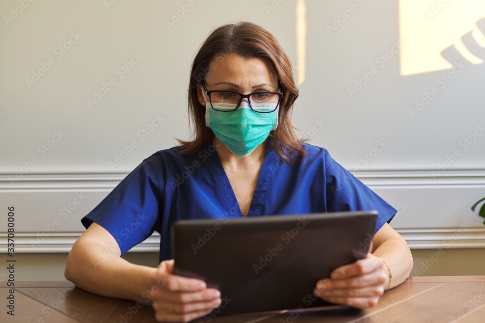 Portrait of doctor woman in medical protective mask with digital tablet sitting at desk in office