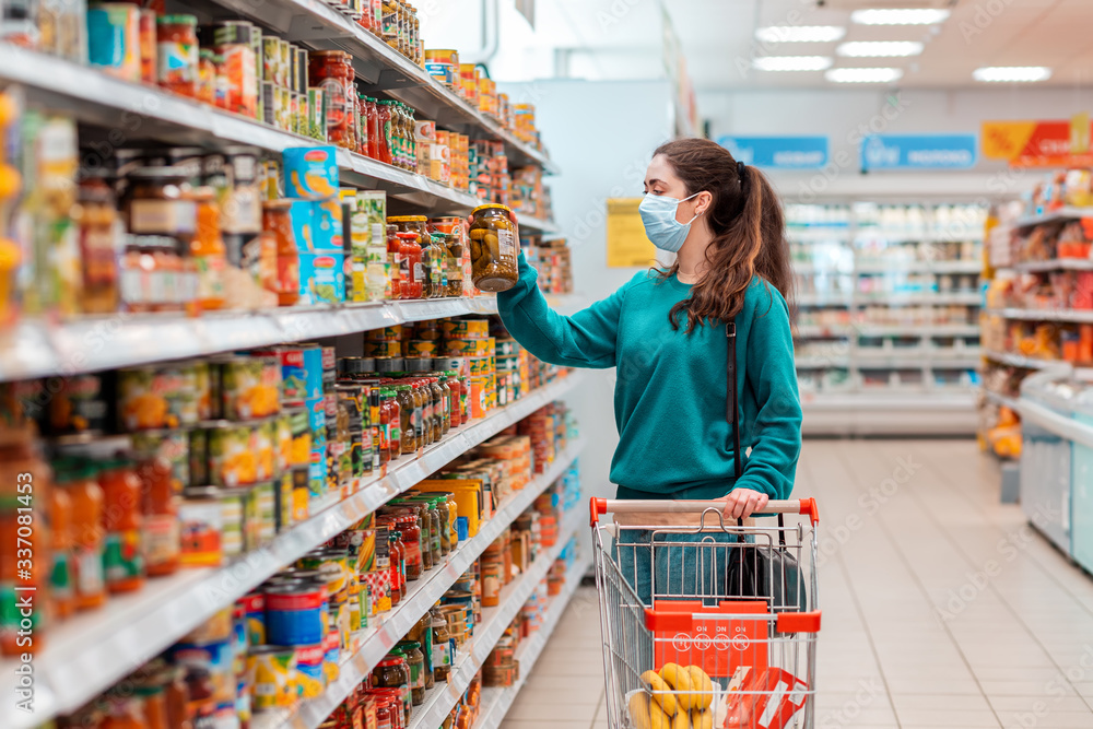 A young pretty Caucasian woman in a medical mask buys a preserved glass jar with vegetable mix at a grocery store. The concept of buying products and the coronovirus pandemic