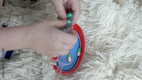 a boy, in an orange mask, wearing glasses, sitting on the floor on a light blanket, squeezes tablets capsules, of white, and green color, from the package into a toy plate for toys in the big plan. photo