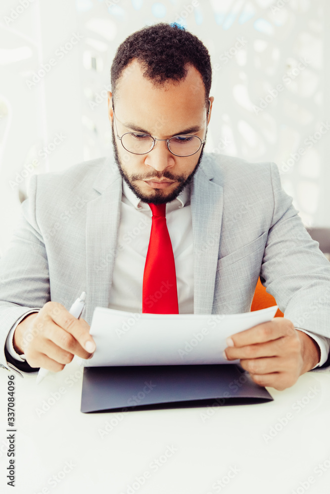 Serious businessman working with contract. Focused young African American businessman in eyeglasses sitting at table and holding documents in office. Paperwork concept Serious businessman working with contract. Focused young African American businessman in eyeglasses sitting at table and holding documents in office. Paperwork concept