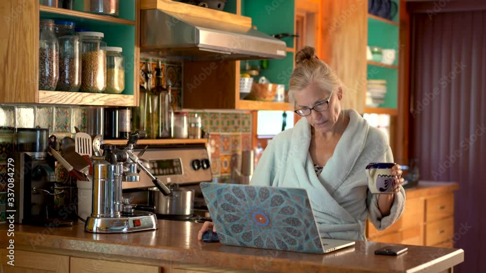 Holding a cup of coffee in one hand and working on a computer with the other, a mature woman in bathrobe nods her head and looks happy in a kitchen setting. Holding a cup of coffee in one hand and working on a computer with the other, a mature woman in bathrobe nods her head and looks happy in a kitchen setting.