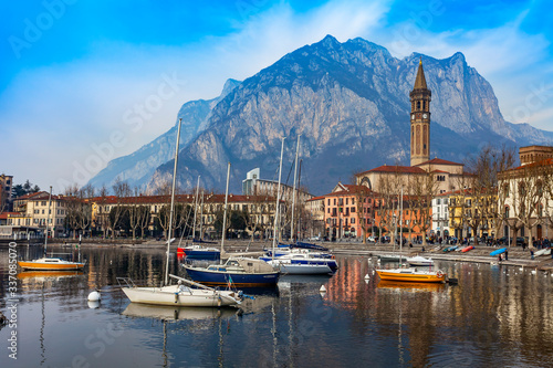 Lecco, Italy, February 16, 2020. The picturesque embankment of Lake Como in cloudy weather and the mountains behind it. Boats are reflected in the water