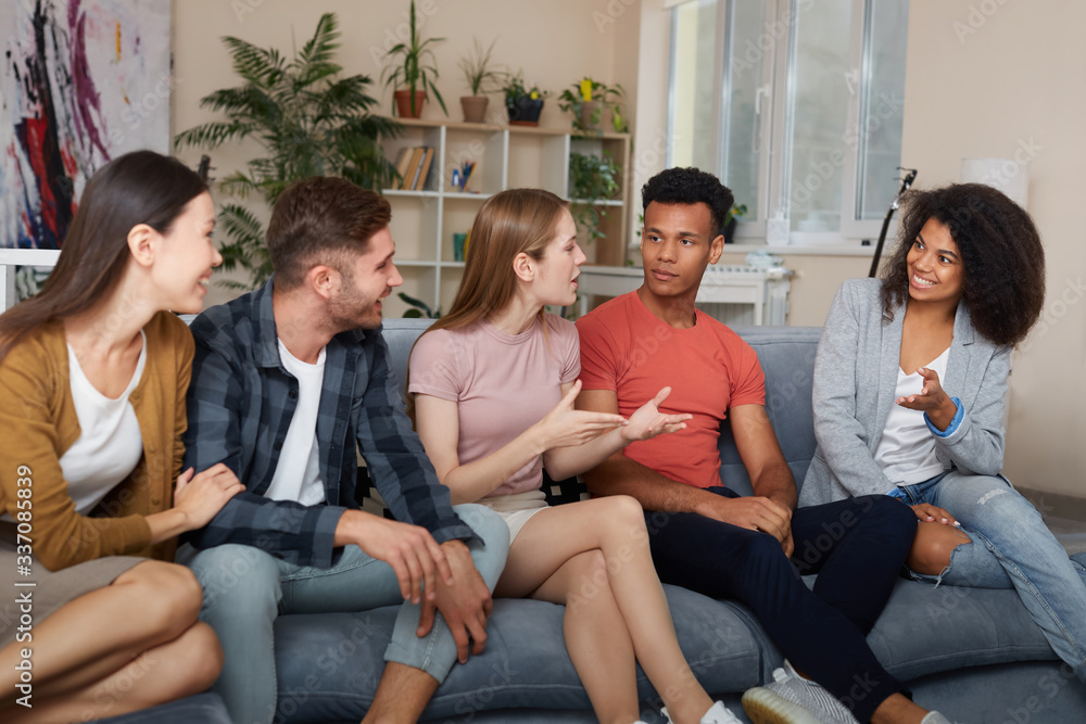Spending time with friends. Group of young multicultural people in casual wear talking and resting together while sitting on the sofa at home. They enjoying time together Spending time with friends. Group of young multicultural people in casual wear talking and resting together while sitting on the sofa at home. They enjoying time together