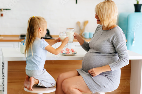 A little girl with mom, a young girl with a pregnant blonde, is poured and drink milk in the kitchen at the table