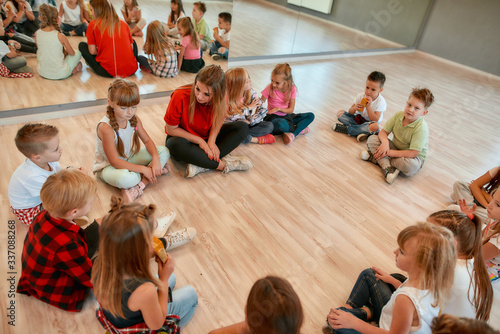 Analyzing mistakes. A group of little dancers sitting on the floor gathered around their female dance teacher and listening her carefully. Dance school