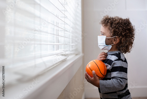A young boy looking out the window holding his basketball wearing a protective facemark while seeking protection from COVID-19, or the novel coronavirus, by sheltering in place in his home. He is read photo