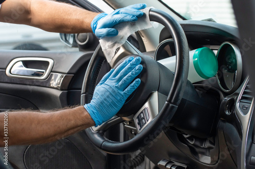 A bearded brown haired man wearing a baseball cap uses alcohol wipes to sterilize the front driver seat, steering wheel, and surrounding areas from any lingering virus strains or germs that are inside