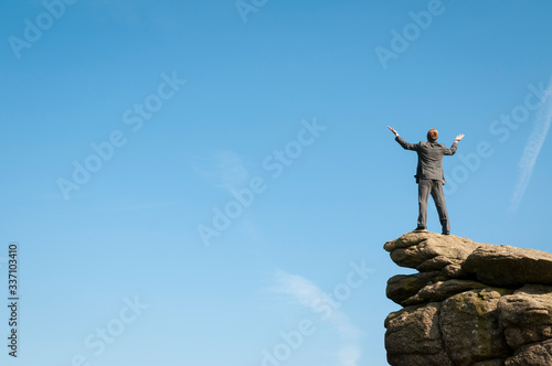 Distant businessman standing with his arms spread on a dramatic rocky mountaintop