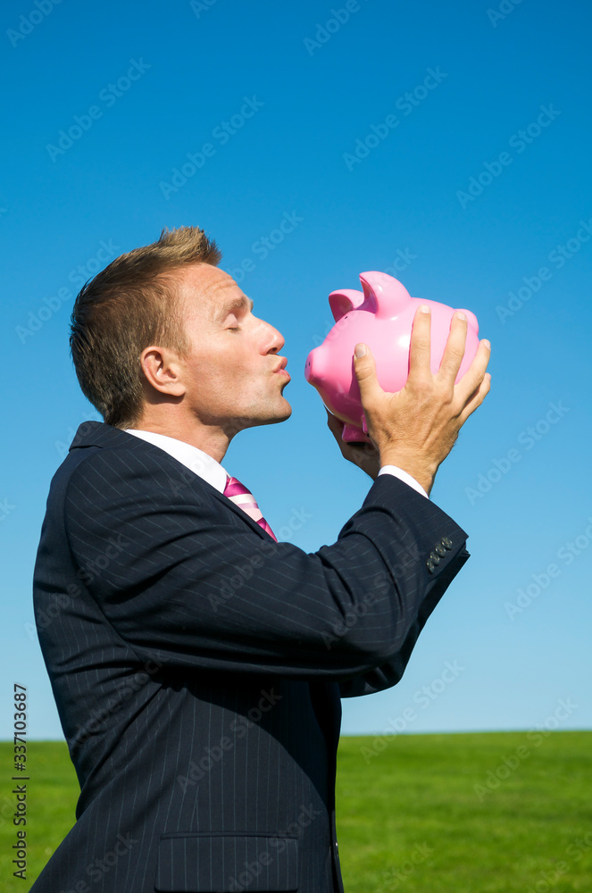Happy businessman kissing pink piggy bank outdoors on bright green meadow with blue sky copy space Happy businessman kissing pink piggy bank outdoors on bright green meadow with blue sky copy space