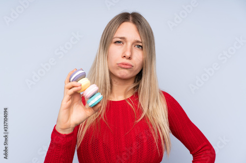Young blonde woman over isolated blue background holding colorful French macarons and with sad expression