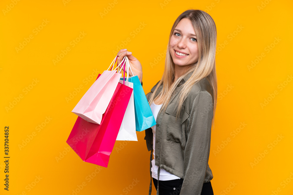Young blonde woman over isolated yellow background holding shopping bags and smiling