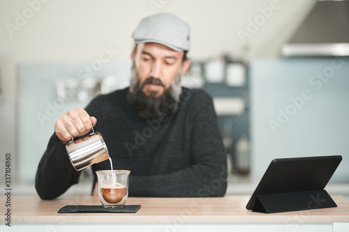 Man pouring milk into coffee on a table with a tablet. Selective focus on the coffee, the man is gently out of focus. Early morning routine concept