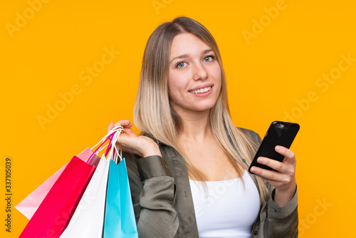 Young blonde woman over isolated yellow background holding shopping bags and writing a message with her cell phone to a friend