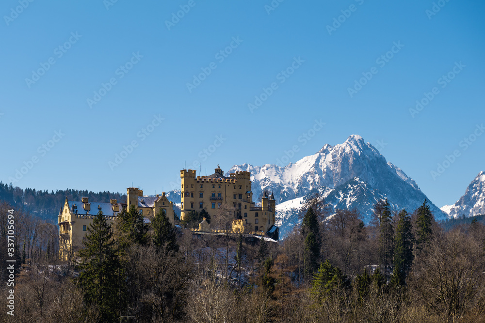 Schloss Hohenschwangau im Frühling mit schneebedeckter Gehrenspitze im Hintergrund vor blauem Himmel