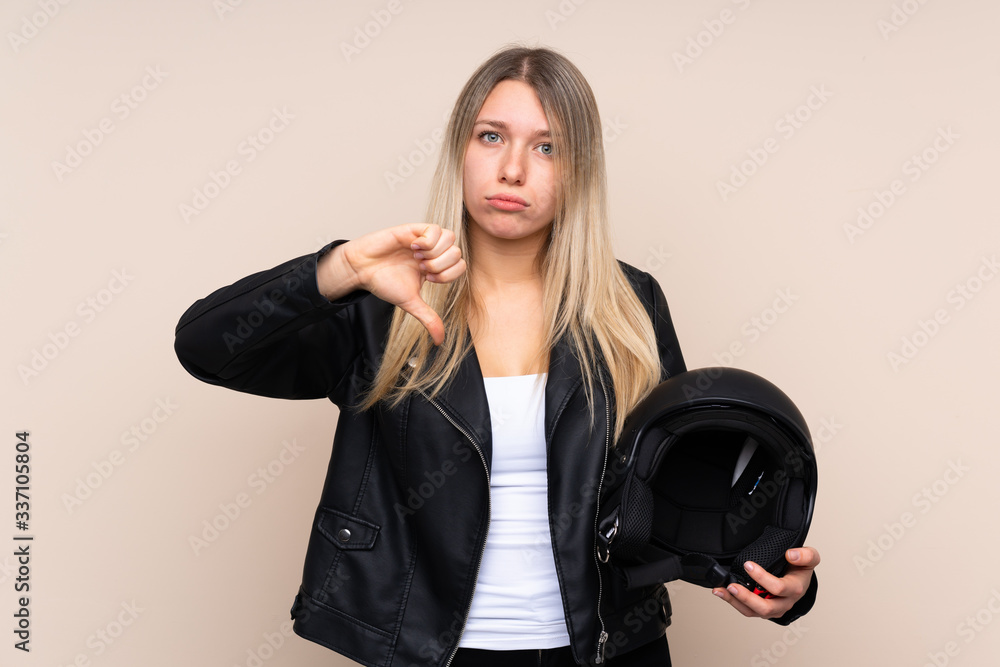 Young blonde woman with a motorcycle helmet over isolated background showing thumb down sign Young blonde woman with a motorcycle helmet over isolated background showing thumb down sign