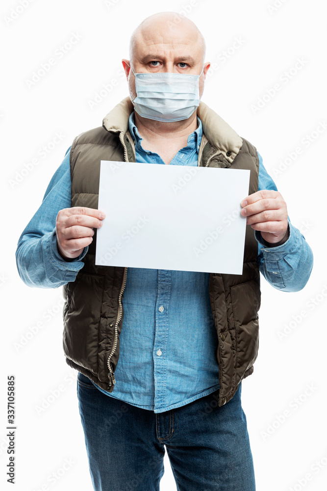 Bald adult man in a medical mask with a blank white sign in his hands. White background. Precautions during the coronavirus pandemic. Vertical. Stay at home. Bald adult man in a medical mask with a blank white sign in his hands. White background. Precautions during the coronavirus pandemic. Vertical. Stay at home.