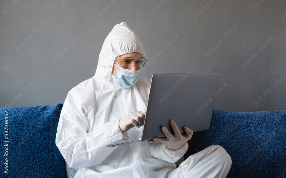 Man in protective suit, medical mask and rubber gloves sits at home and works with laptop at the table during quarantine. Designer, artist, architect, businessman at remote work in a pandemic covid. Man in protective suit, medical mask and rubber gloves sits at home and works with laptop at the table during quarantine. Designer, artist, architect, businessman at remote work in a pandemic covid.