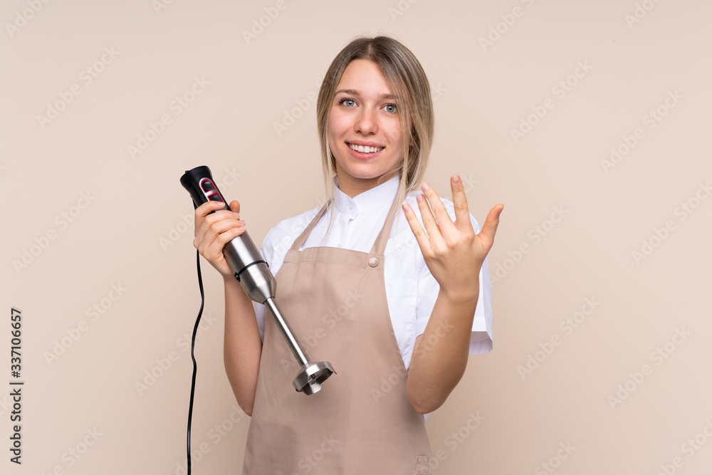 Young blonde woman using hand blender inviting to come with hand. Happy that you came Young blonde woman using hand blender inviting to come with hand. Happy that you came
