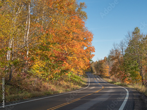 Colorful yellow and orange leaves line a two-lane road winding through the northern woods on a sunny fall day in the United States.