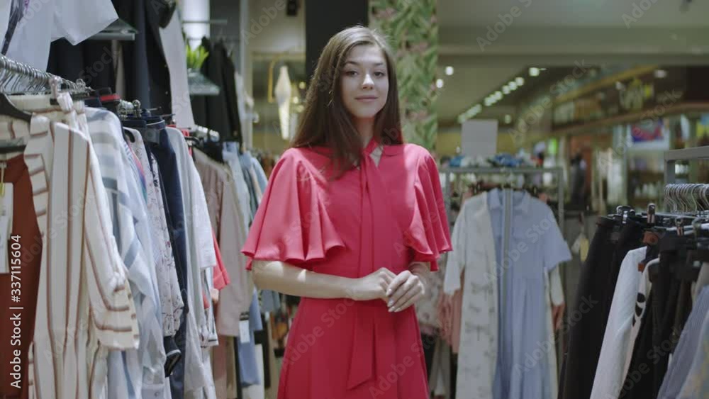 Smiling attractive girl is holding and crossing her arms while posing for the camera in clothes shop. Front view of beautiful brunette woman standing between clothes racks in the mall. Smiling attractive girl is holding and crossing her arms while posing for the camera in clothes shop. Front view of beautiful brunette woman standing between clothes racks in the mall.