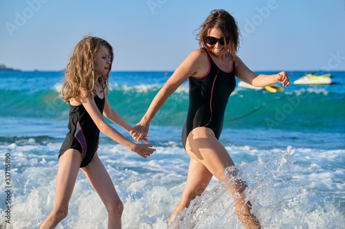 Mother and daughter child in swimsuits walking along seashore holding hands