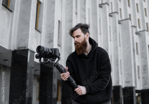 Bearded Professional videographer in black hoodie holding professional camera on 3-axis gimbal stabilizer. Filmmaker making a great video with a professional cinema camera. Cinematographer.