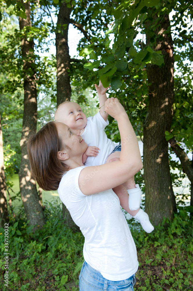 Young woman with a one-year-old baby in her arms in a white T-shirt in the park smiles and looks at the leaves of the trees Mother's Day Young woman with a one-year-old baby in her arms in a white T-shirt in the park smiles and looks at the leaves of the trees Mother's Day