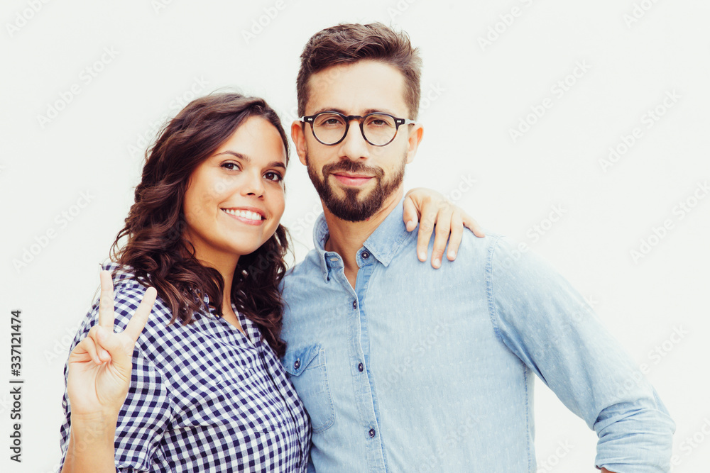Cheerful beautiful woman hugging boyfriend and making peace sign. Young woman in casual and man in glasses in glasses posing isolated over white background. Happy relationship concept Cheerful beautiful woman hugging boyfriend and making peace sign. Young woman in casual and man in glasses in glasses posing isolated over white background. Happy relationship concept